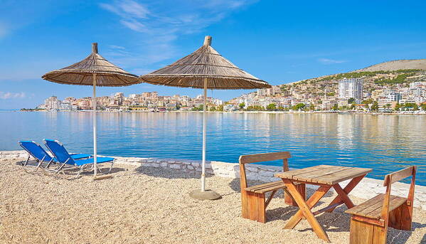 Peaceful beach with umbrellas, chairs, and a view of Saranda across the bay.